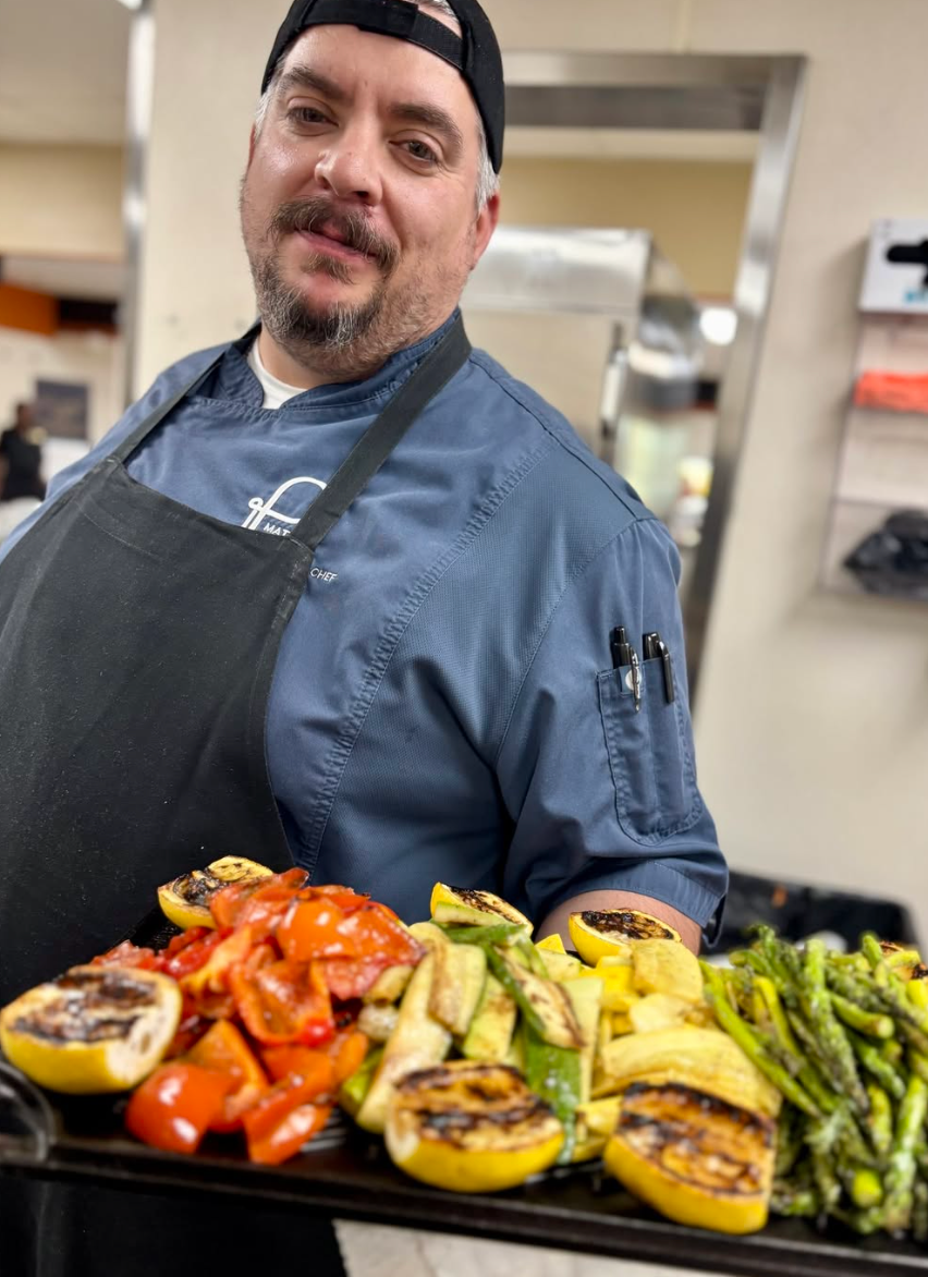 Chef holding a platter of grilled vegetables, including peppers, zucchini, and asparagus. He smiles at the camera.