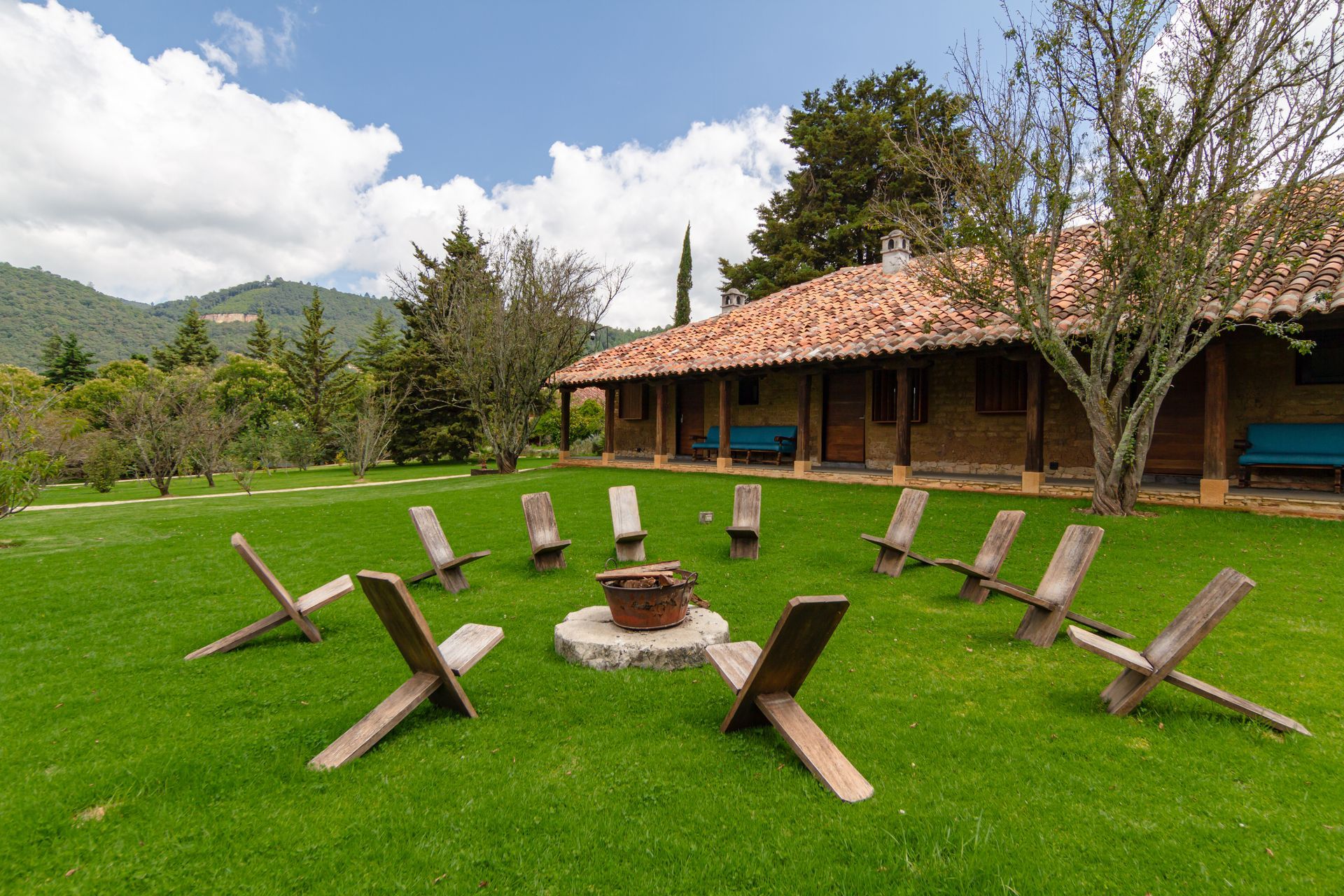 A group of wooden chairs are sitting around a fire pit in the grass in front of a house.
