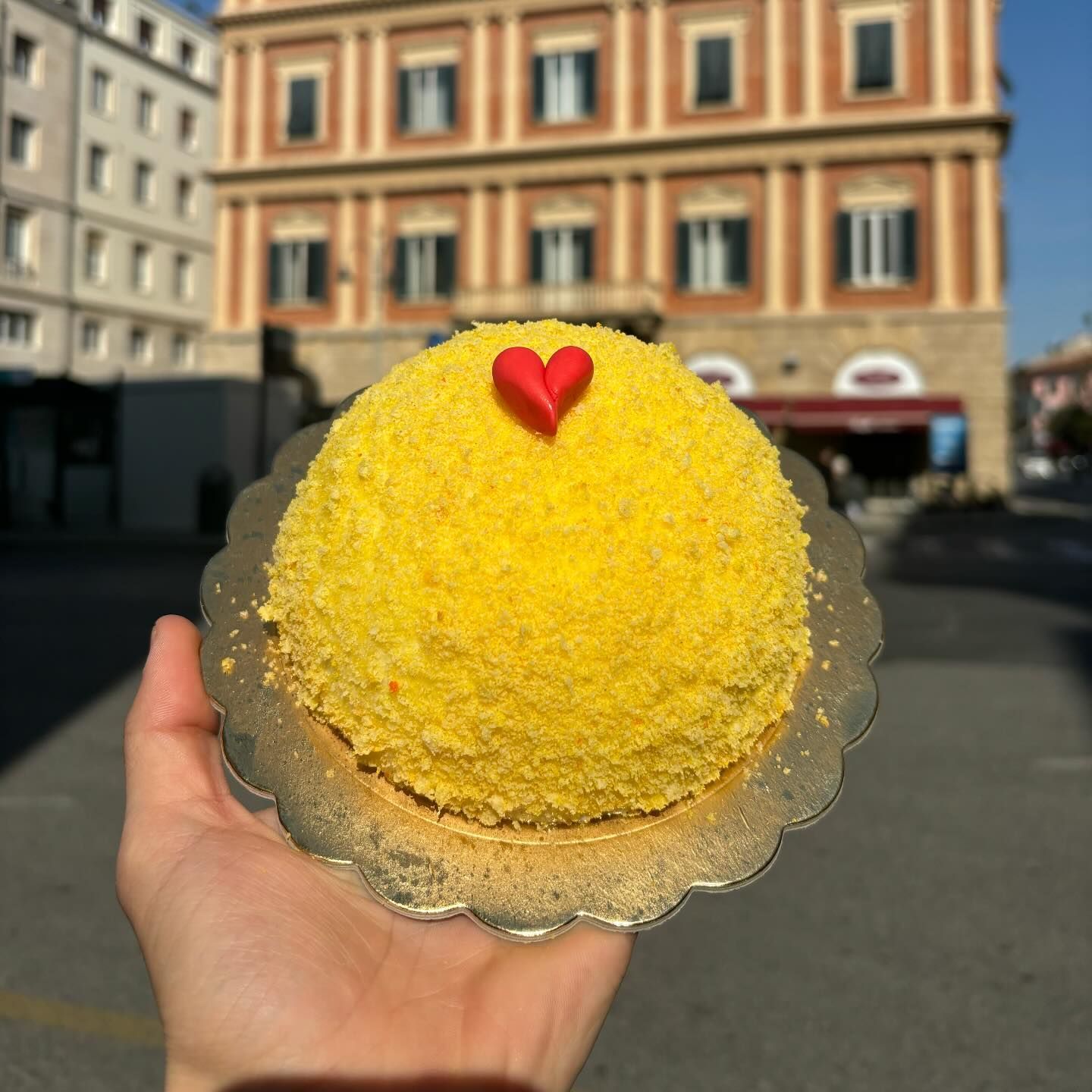 Mano che regge una torta gialla con un cuore rosso su un piatto d'argento, di fronte a un edificio.