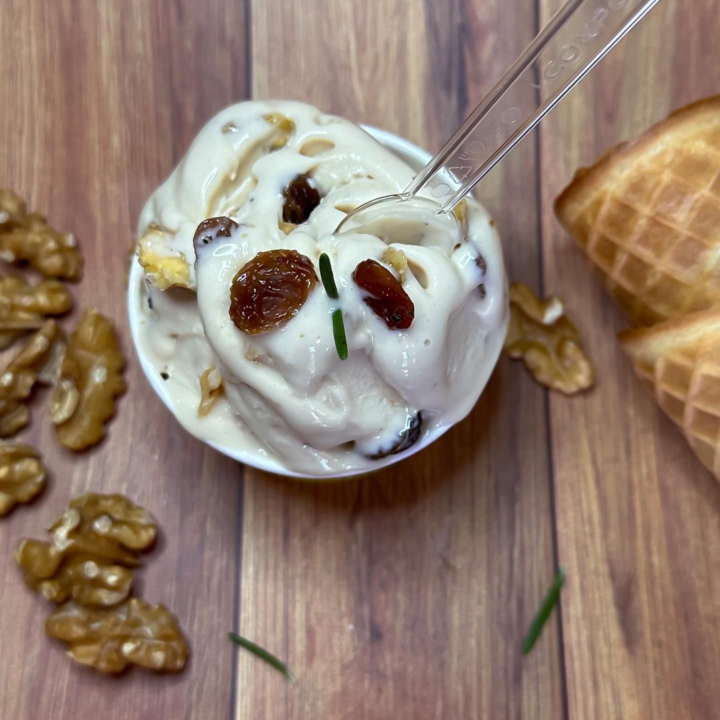 Gelato con noci e uvetta in una coppetta bianca, un cucchiaio, coni di cialda, su un tavolo di legno.