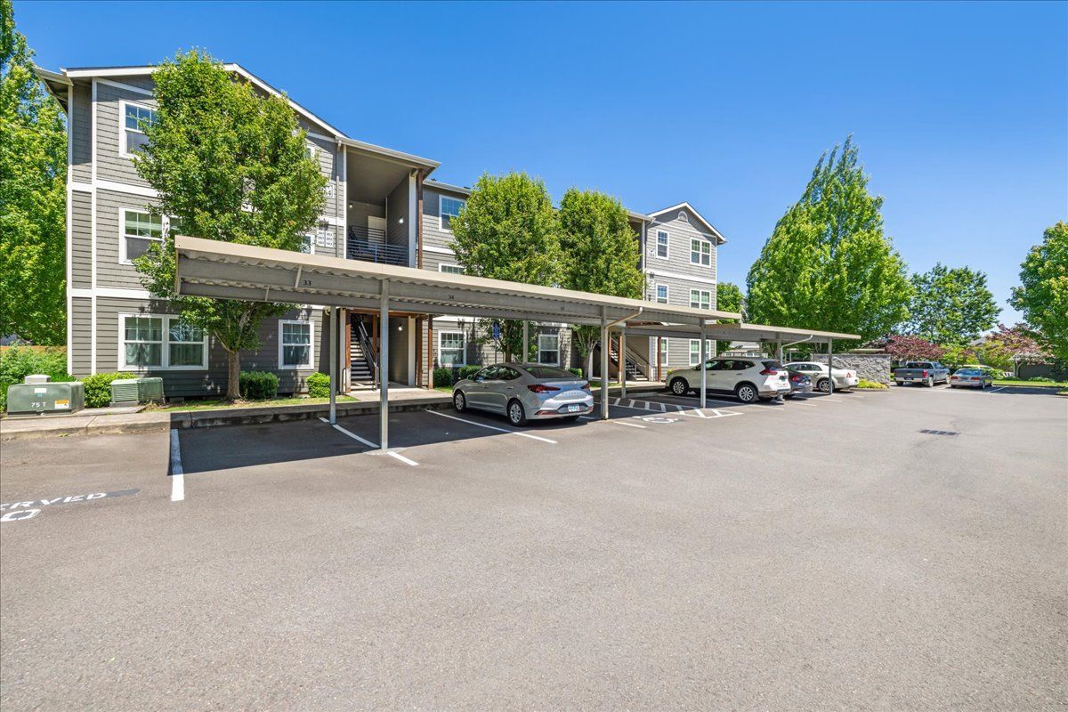 Apartment building with covered parking. Gray siding, blue sky, cars parked under metal canopies.
