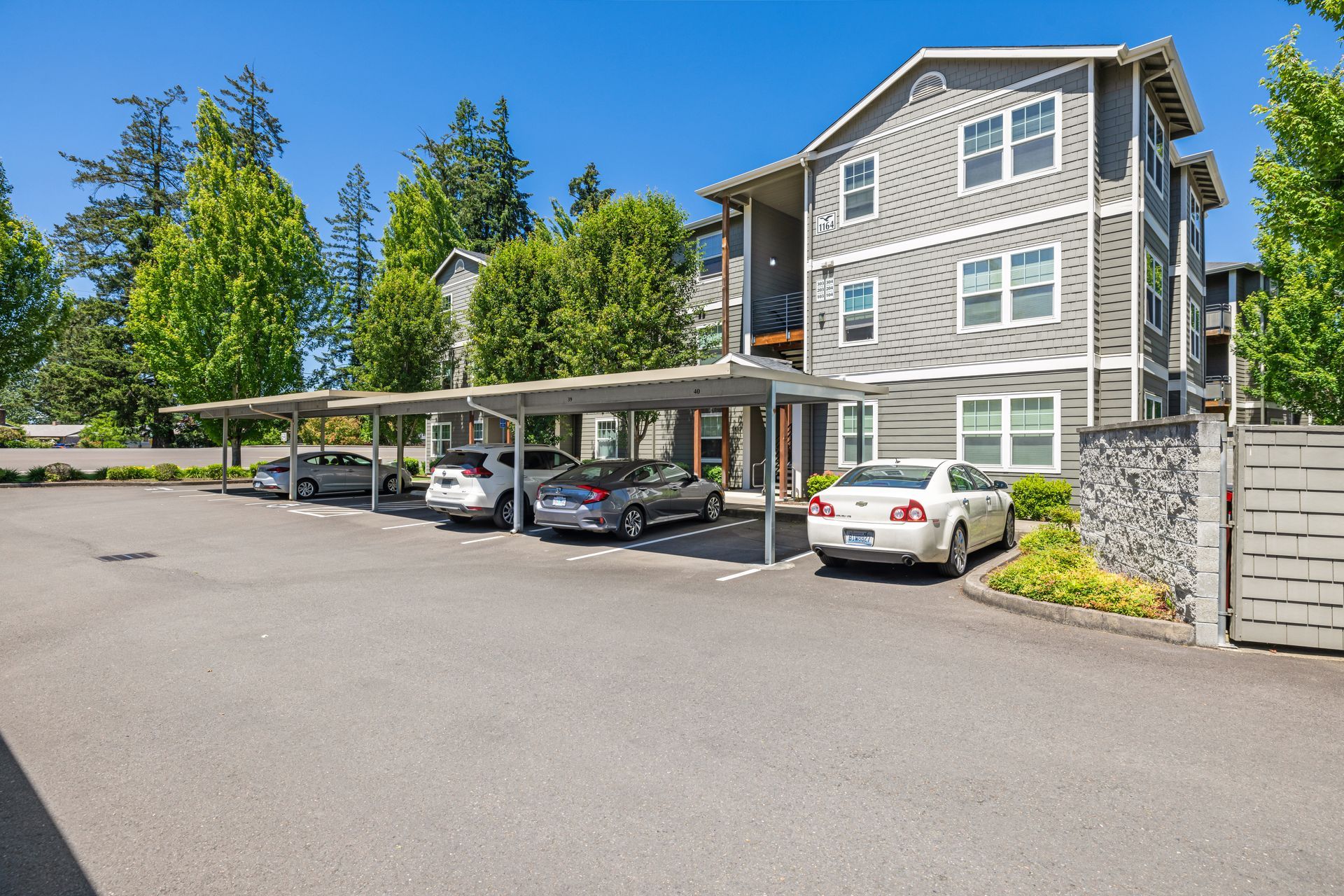 Apartment building with covered parking. Several cars parked under the canopy. Blue sky and trees in the background.