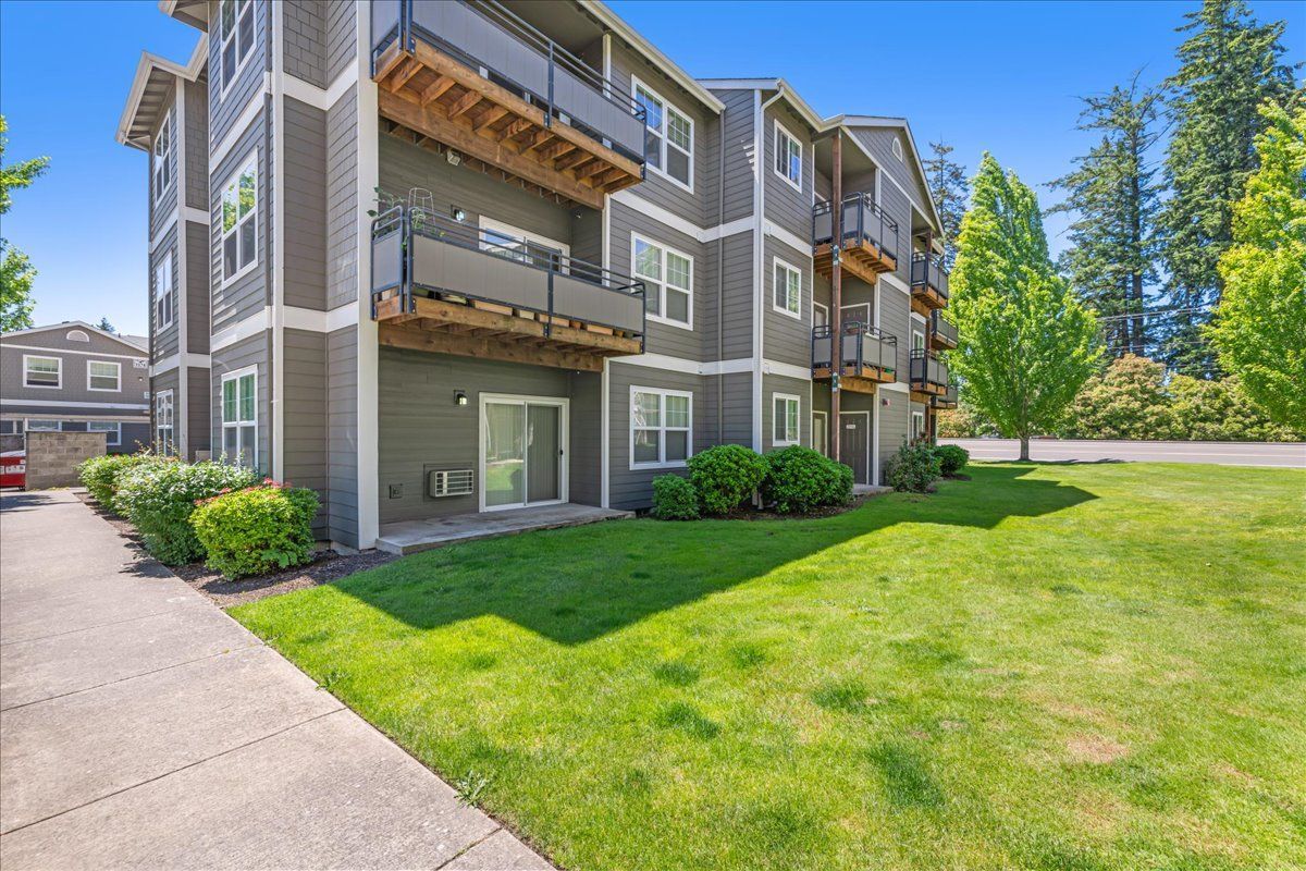 Multi-story apartment building with balconies, green lawn, and sidewalk on a sunny day.
