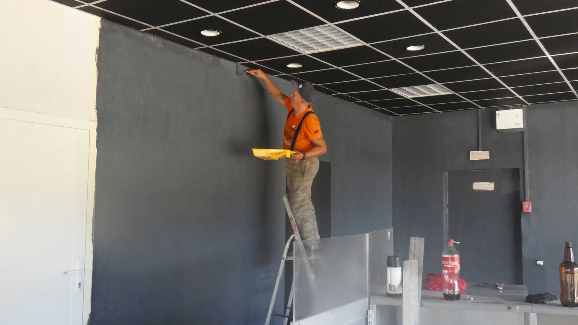 Worker on a step ladder installing ceiling tiles in a room with dark walls and a black grid ceiling.
