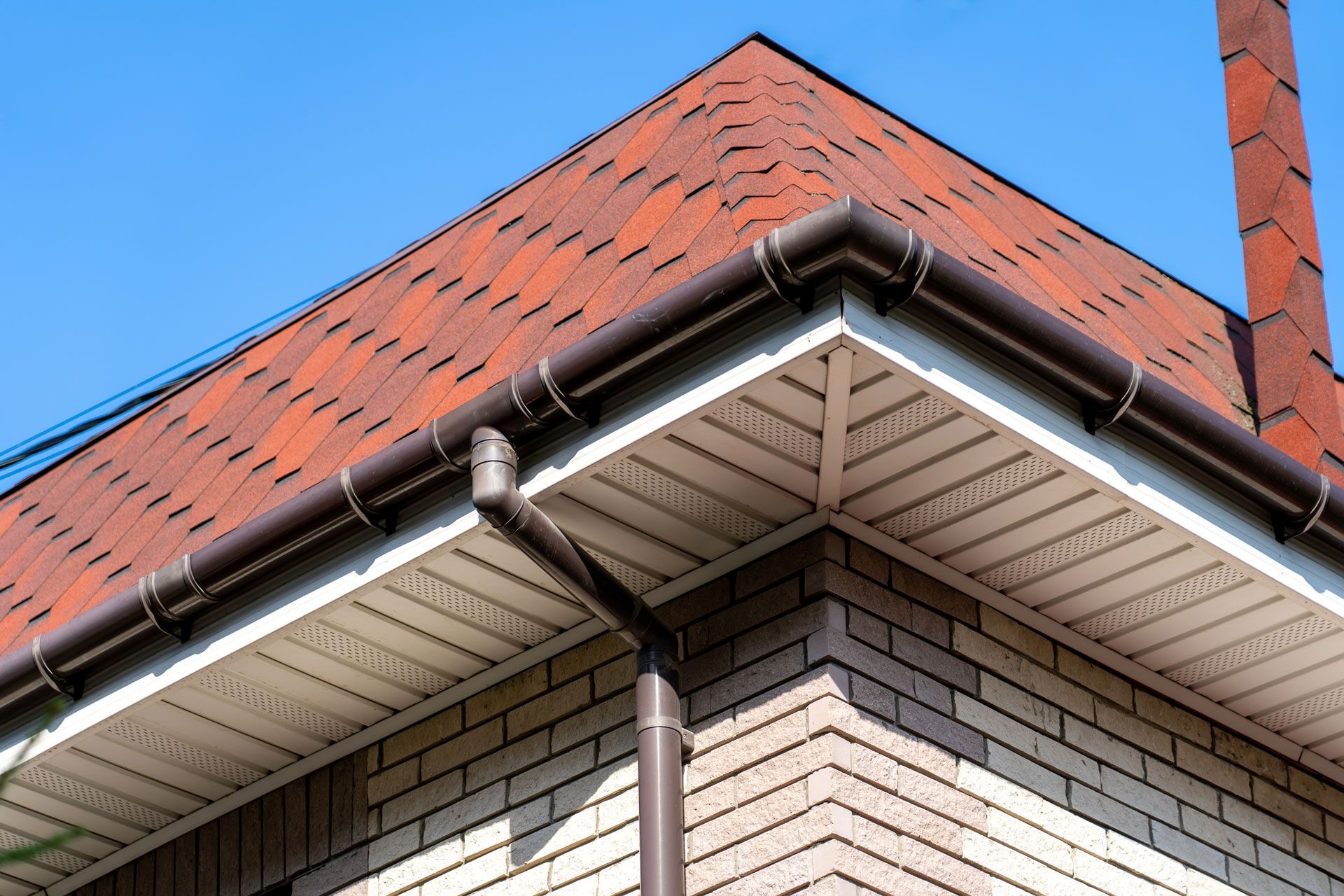 Close-up of a red-tiled roof corner with white gutters and a brick chimney against a blue sky