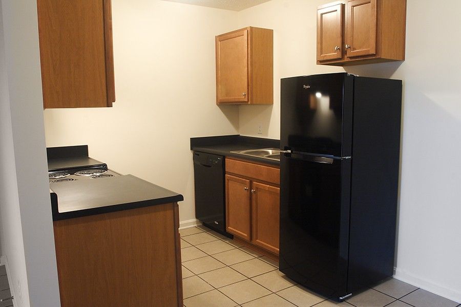 A small kitchen with light-colored walls, wood cabinets, a black refrigerator, dishwasher, and stove on tiled flooring.
