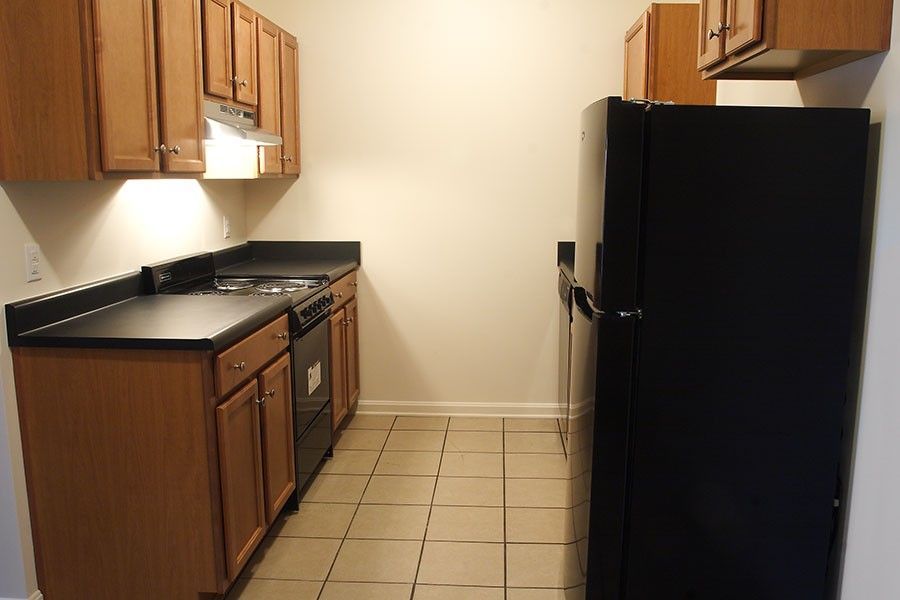 A narrow galley kitchen with light wood cabinets, black countertops, a black refrigerator, and tan floor tiles.