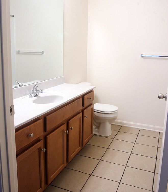 A bathroom with a wooden vanity, a white countertop with a sink, a large mirror, a toilet, and beige tiled flooring.