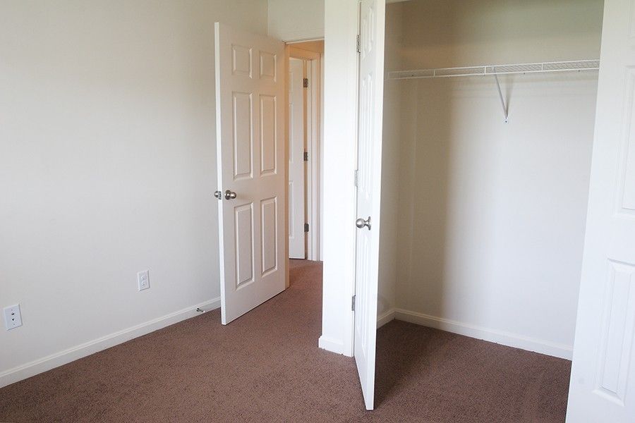 An empty room with tan carpet, white walls, an open bedroom door, and a closet with a wire shelf.