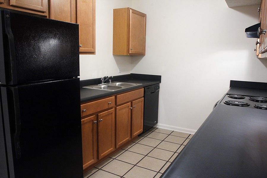 A galley-style kitchen with medium-wood cabinets, black countertops, a black refrigerator, and light-colored tile floors.