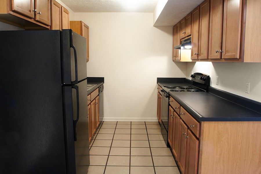 Galley kitchen with light wood cabinets, black countertops, black appliances, and light-colored square floor tiles.