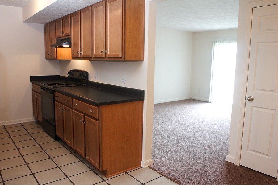 A kitchen with wood cabinets and black counters next to an open living area with carpeted floors and a sliding glass door.