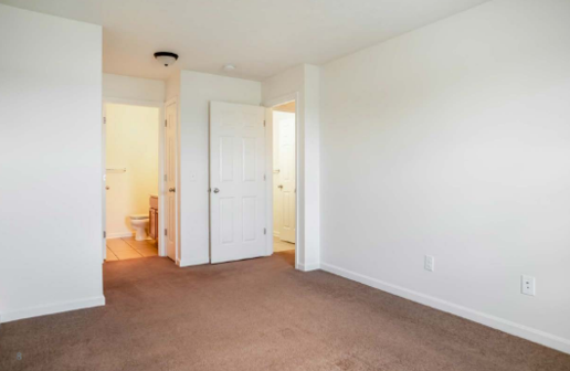 An empty room with beige carpet, white walls, a doorway to a bathroom, and a closed door leading to a closet.