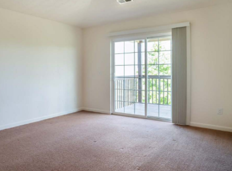 A bright, empty room with plain off-white walls, brown carpet, and a large sliding glass door leading to a balcony.