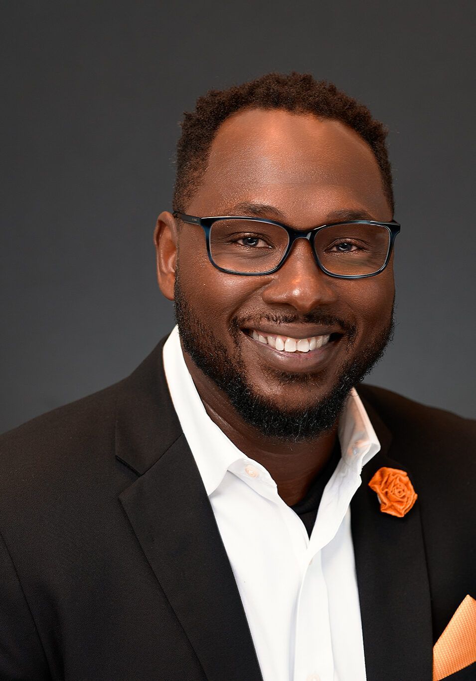 Man in glasses, smiling, wearing a black blazer, white shirt, orange boutonniere, and pocket square.