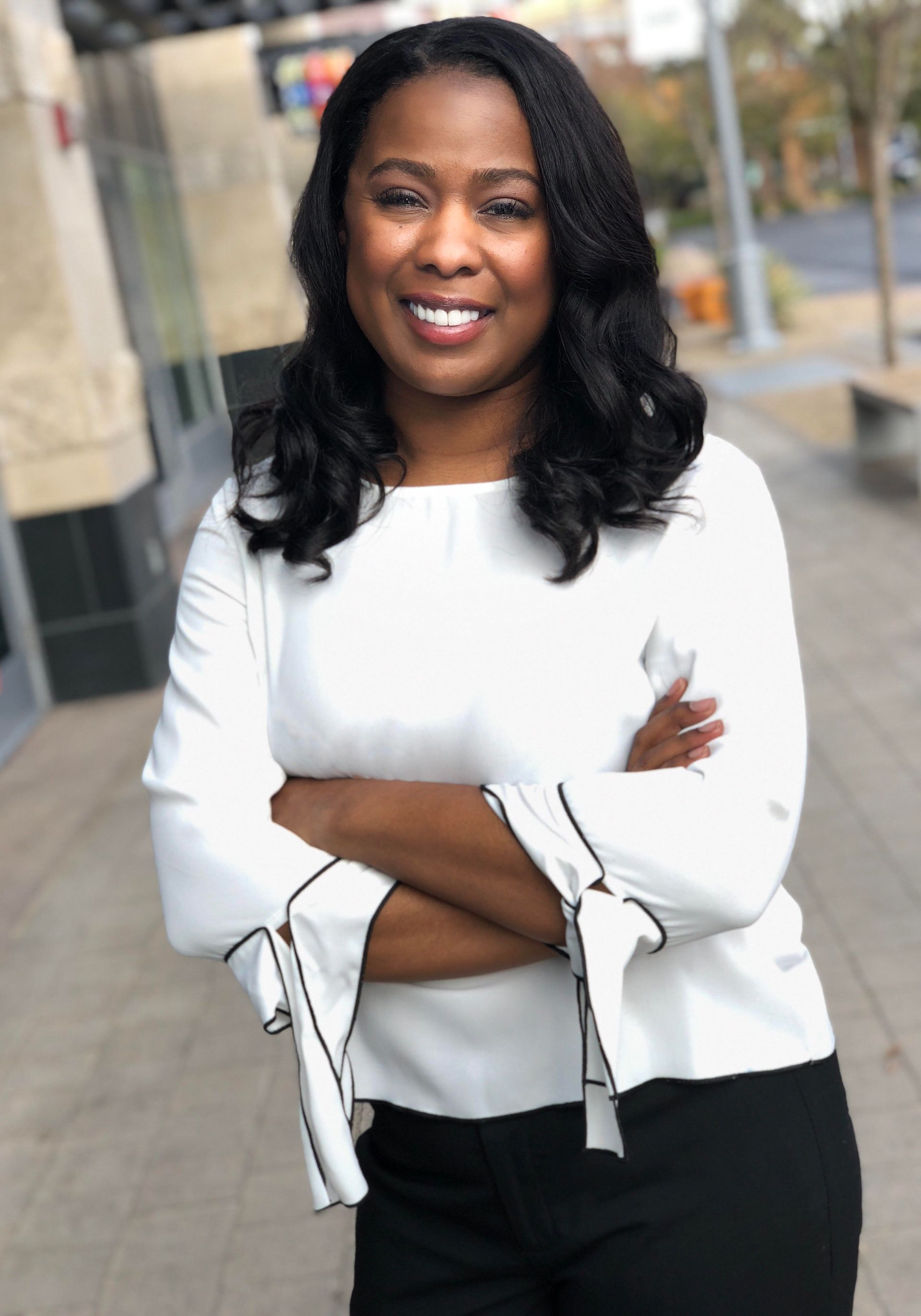 Woman in white top, arms crossed, smiling. Outdoors, urban setting, sunlight.