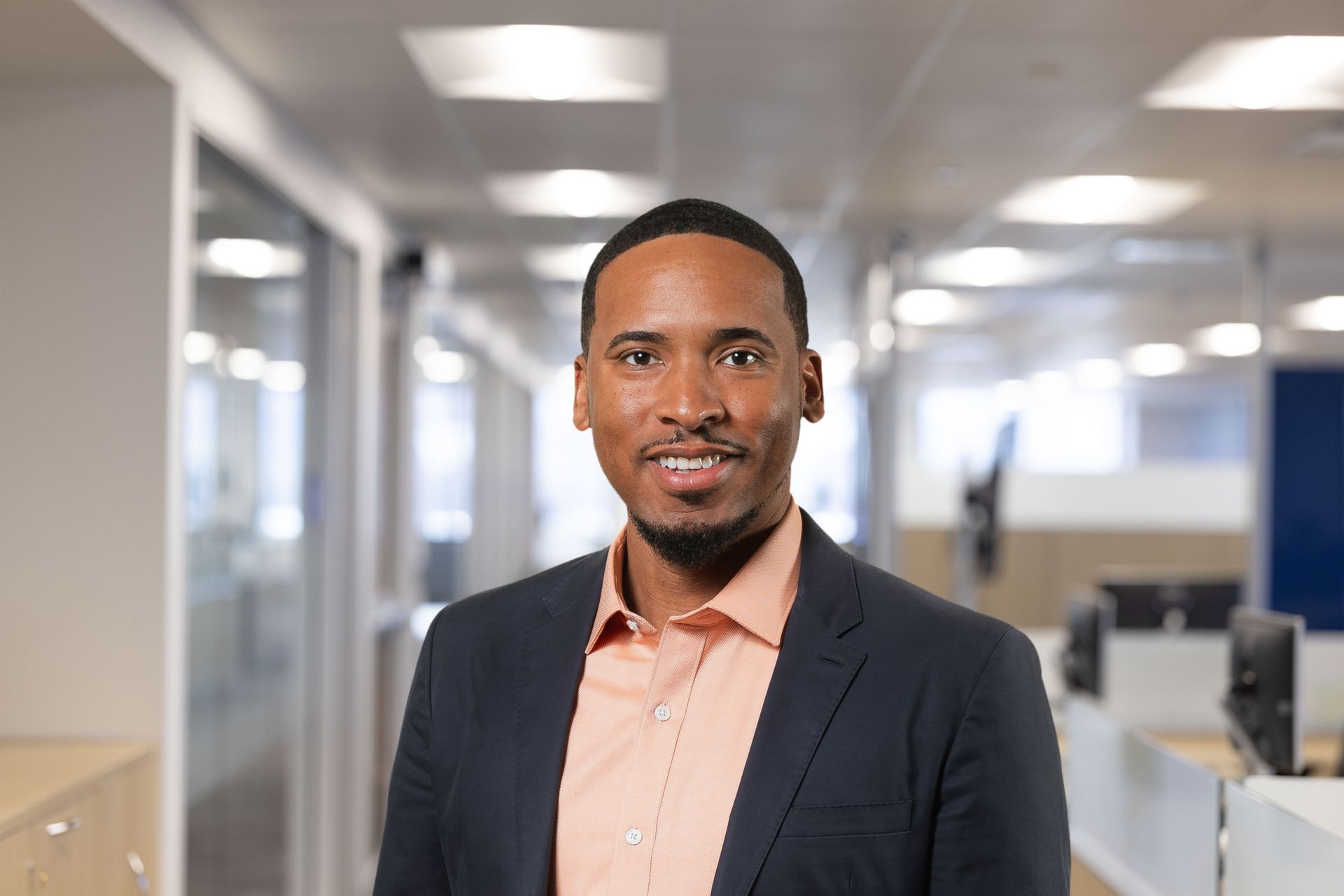 Man in a suit smiles, wearing a tie with a landscape design, standing against a dark background.