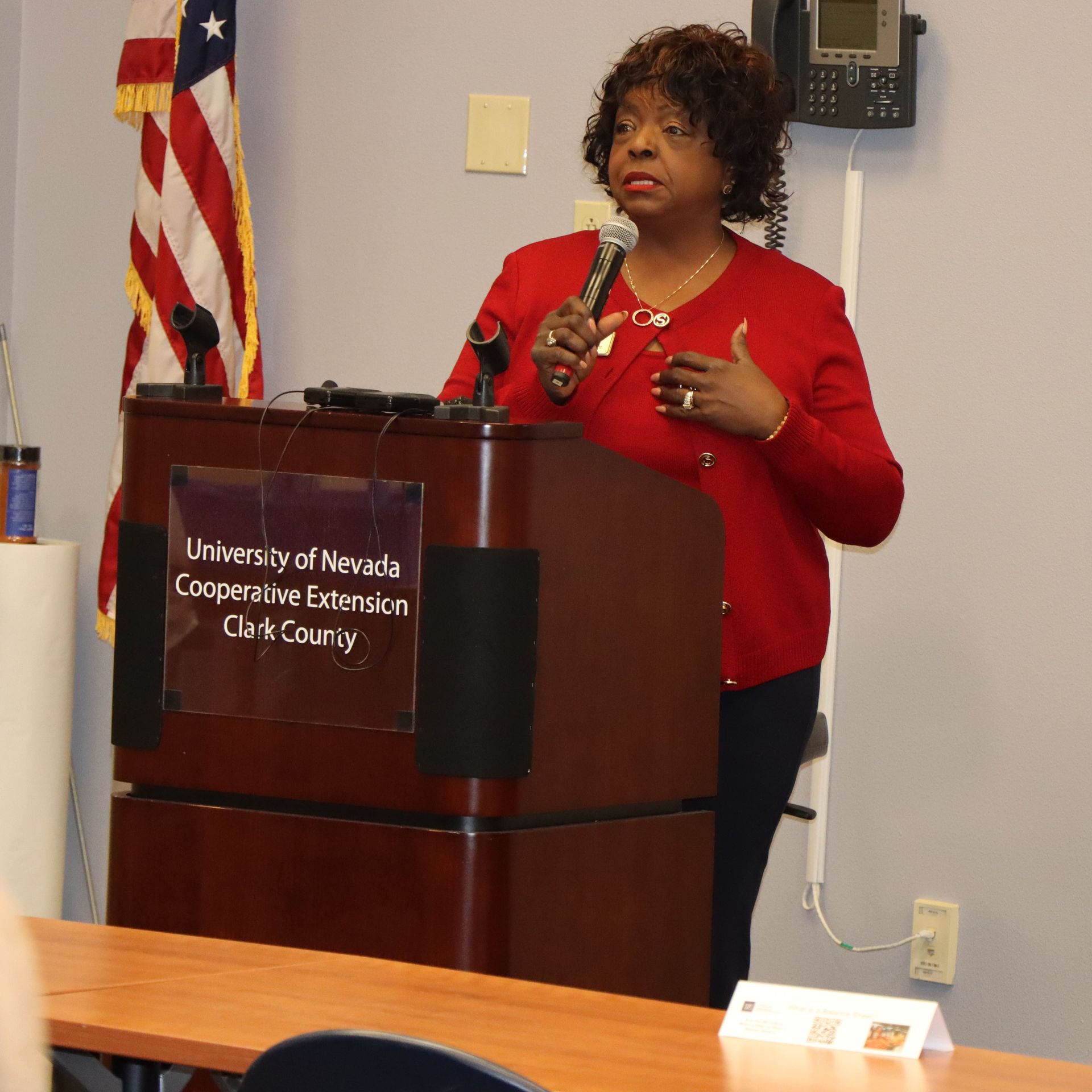 Woman speaking at a podium in an office, holding a microphone. American flag in background.