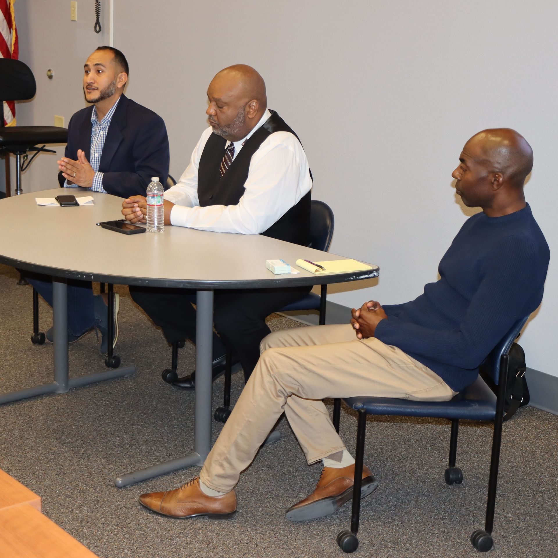 Three men seated at a table. One speaks, another listens intently, the third looks on.