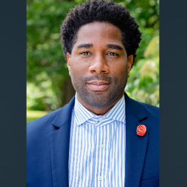 Man in blue suit, striped shirt, red flower pin, looking at the viewer. Blurred outdoor background.