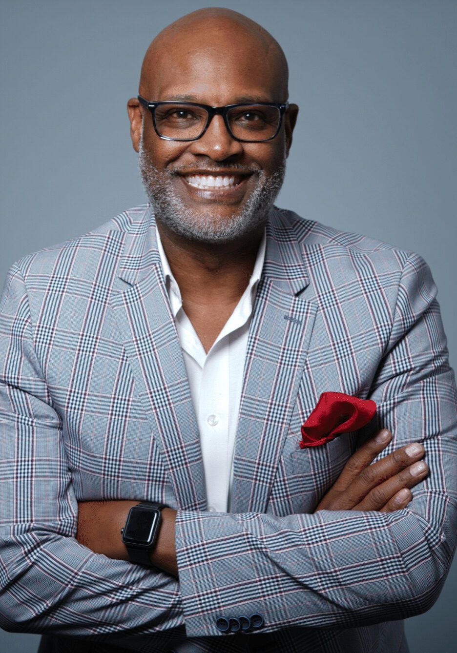 Bald man with glasses smiles, arms crossed, wearing a plaid blazer and white shirt. Pocket square.