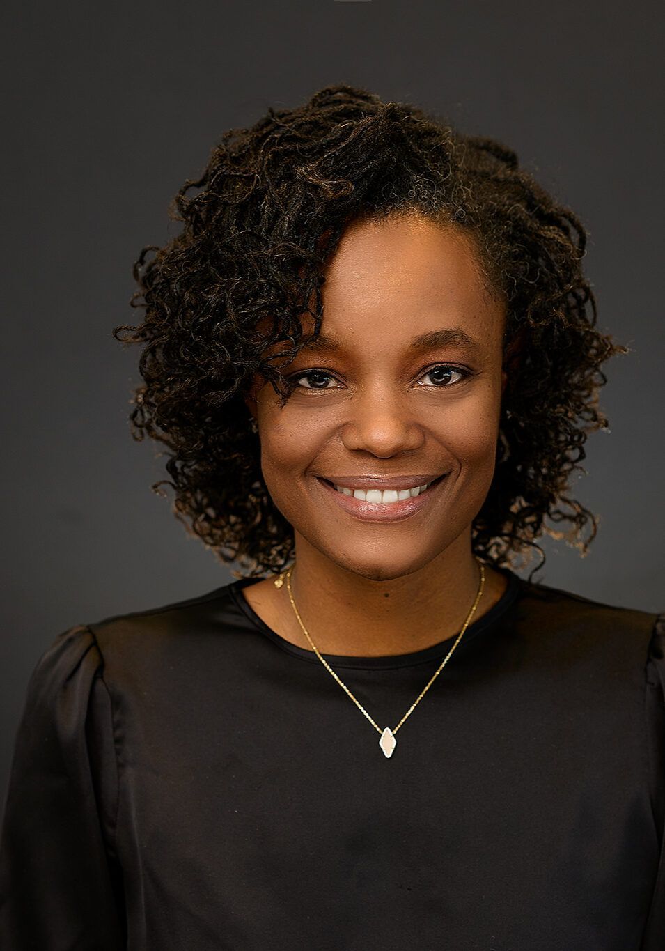 Woman with curly hair smiles, wearing a black shirt and gold necklace, against a gray background.