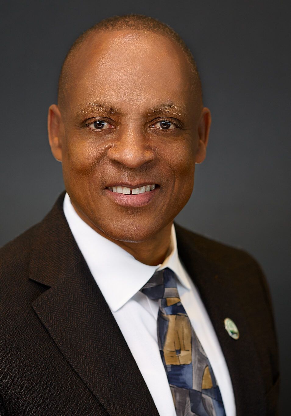 Man in a suit smiles, wearing a tie with a landscape design, standing against a dark background.