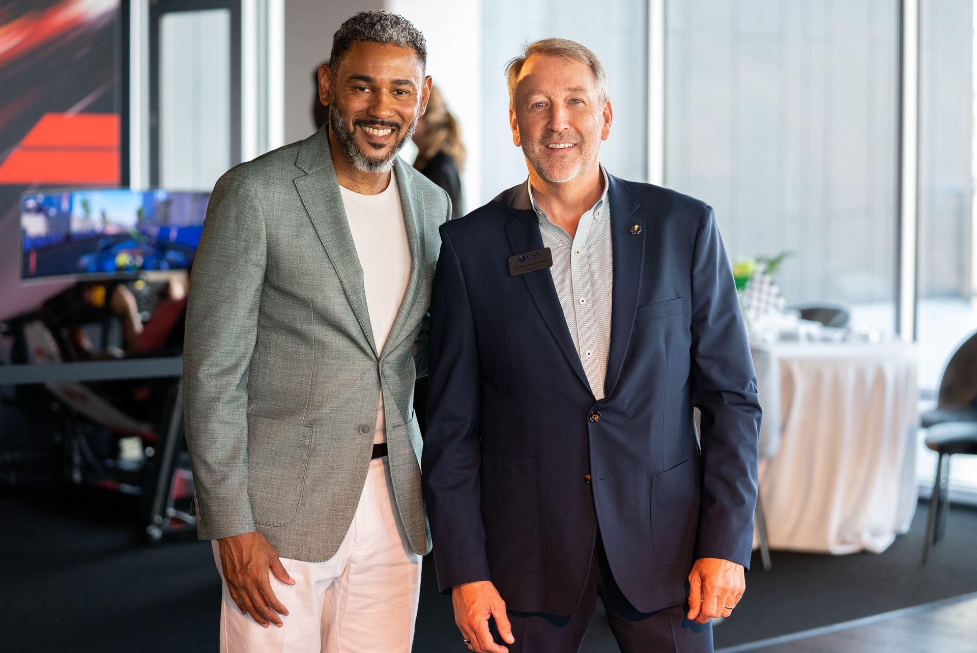 Two men smiling, posing together indoors. One in a gray blazer and white pants, the other in a navy suit.