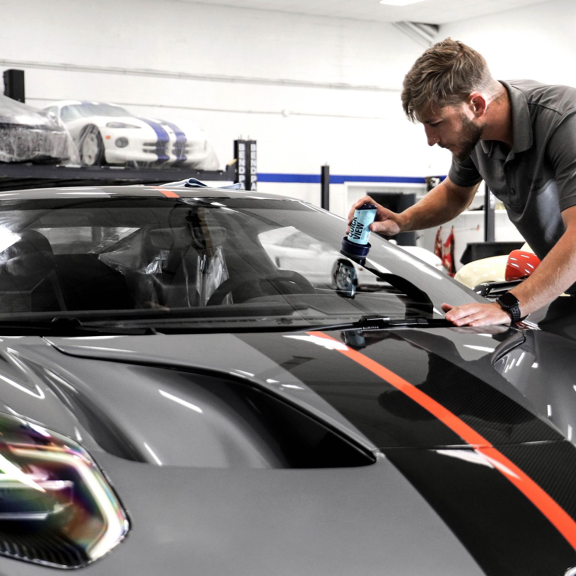 Man applying film to the windshield of a gray sports car in a garage, with orange stripe.