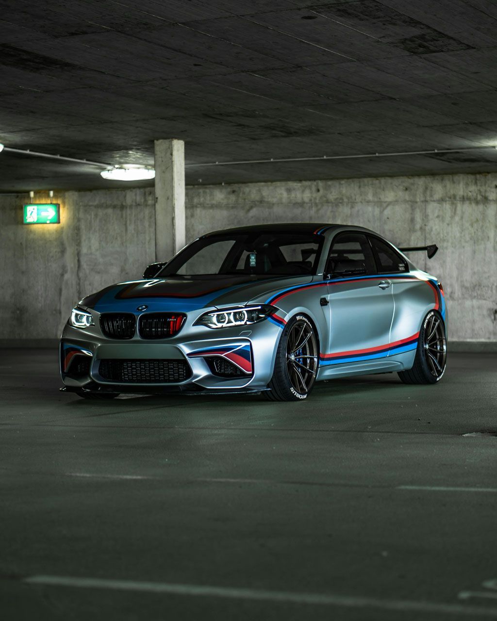 Silver BMW M2 sports car with blue and red stripes in a parking garage.