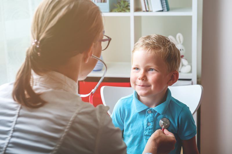 Doctor listening to a young child's chest with a stethoscope in a brightly lit room.