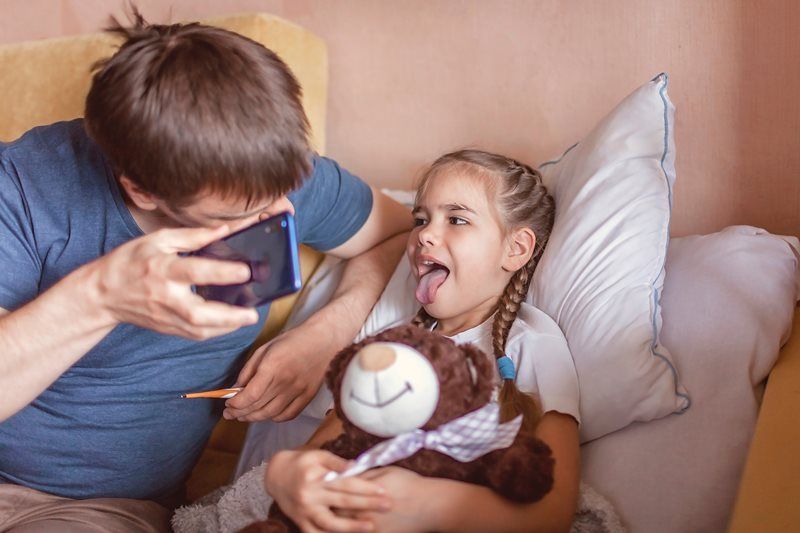 Man examining girl's throat with a smartphone, teddy bear present. Girl lies in bed with mouth open.