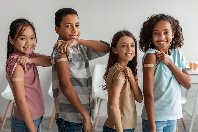 Four children smiling, each with a bandage on their arm, likely after a vaccination. Indoors, white background.