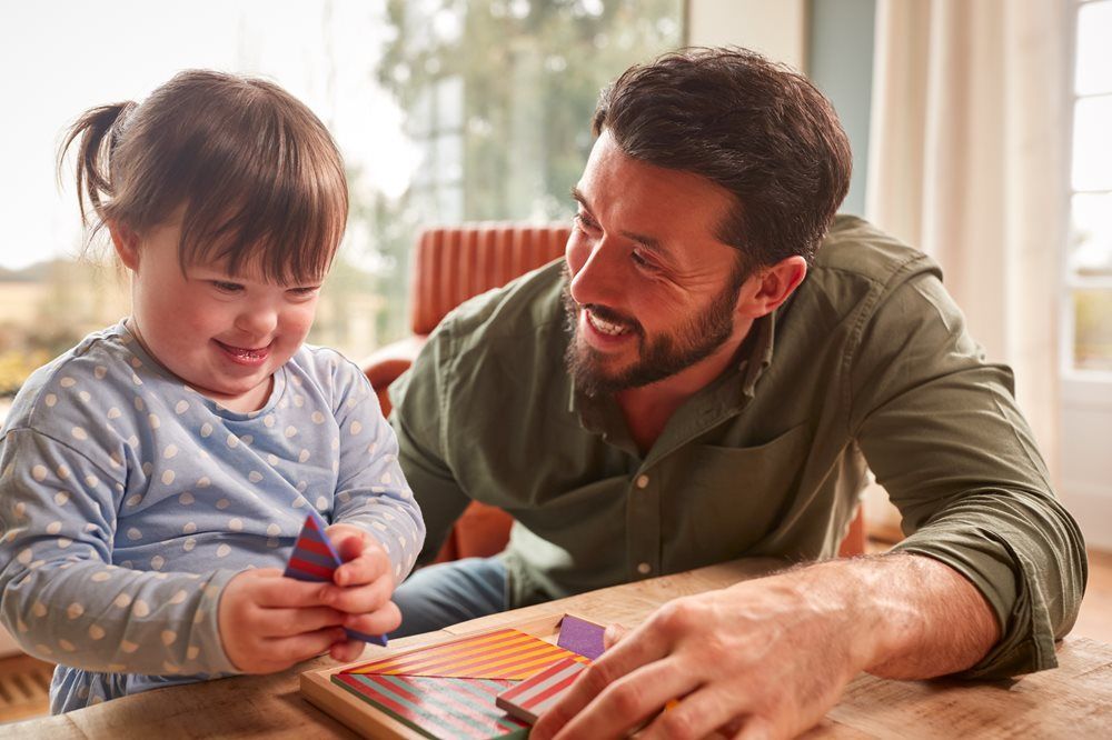 A smiling child with Down syndrome and a man work on a wooden puzzle at a table.