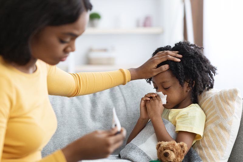 Woman checking child's forehead for fever. Child with tissue, wearing yellow, sitting on couch.
