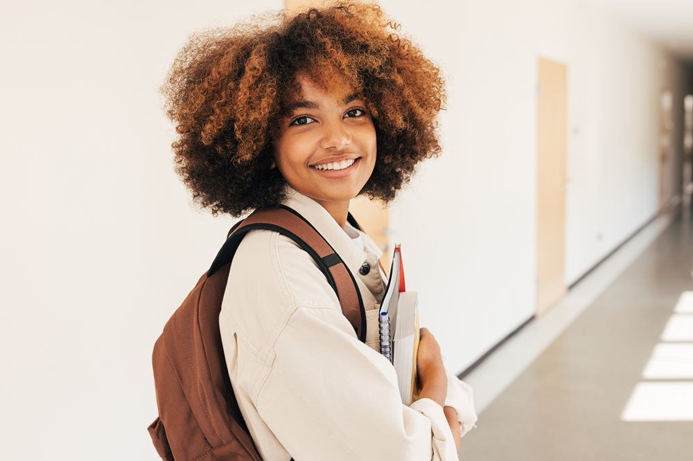Young person with curly brown hair, backpack, and books smiling in a school hallway.