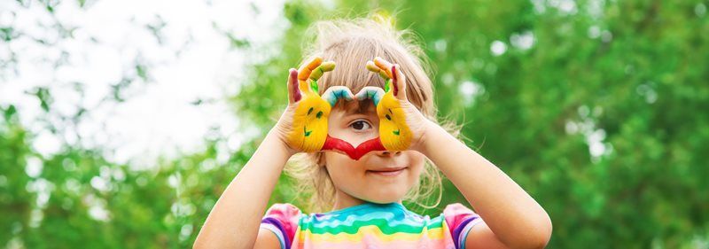 Child with hands painted yellow and red, forming heart shapes over eyes, smiling. Outdoors, green background.