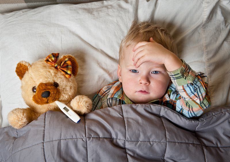 Sick baby in bed, holding forehead. Teddy bear and thermometer nearby.