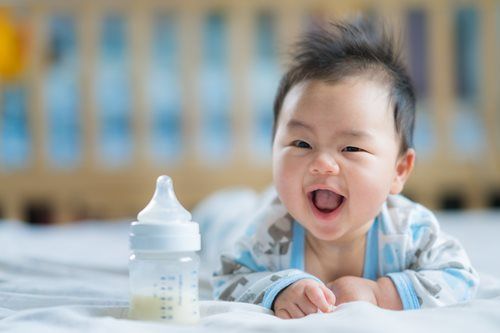 Happy baby with bottle, lying on a white surface, smiling with mouth open.