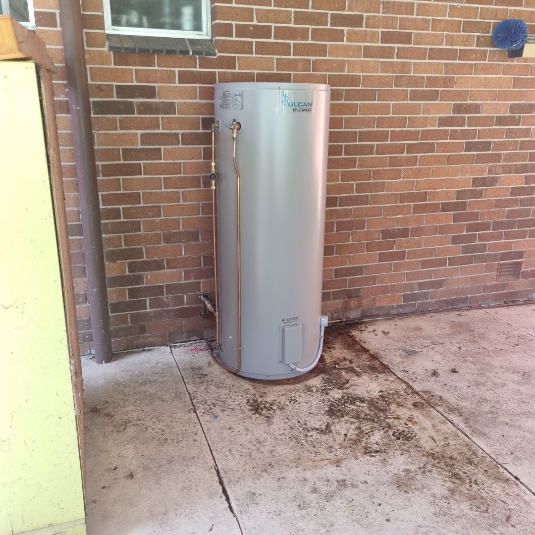A Cylindrical Hot Water Heater Stands Against a Red Brick Wall — Hunter Plumbing And Hotwater in Fern Bay, NSW
