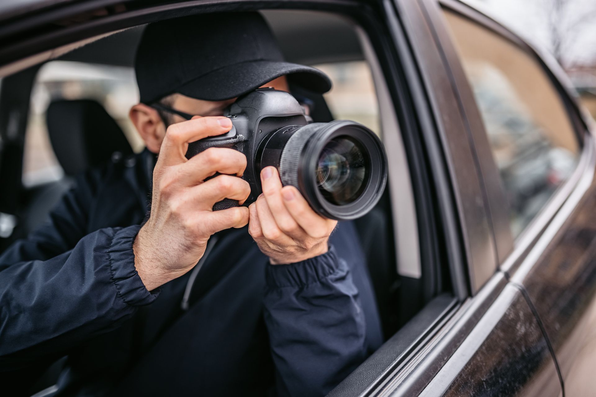 A man wearing a fedora hat and a jacket takes photographs from the window of his white car.