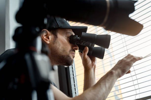 Person looking through binoculars while peeking through window blinds.