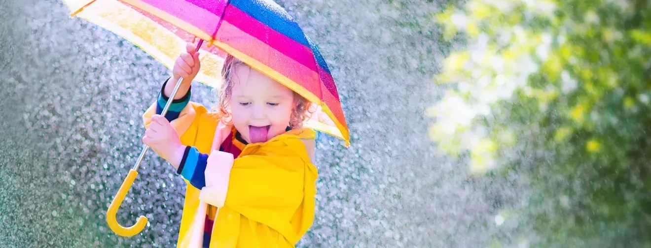 A young girl in a yellow rain jacket is holding a colorful umbrella and smiling. She is standing outside in the rain.