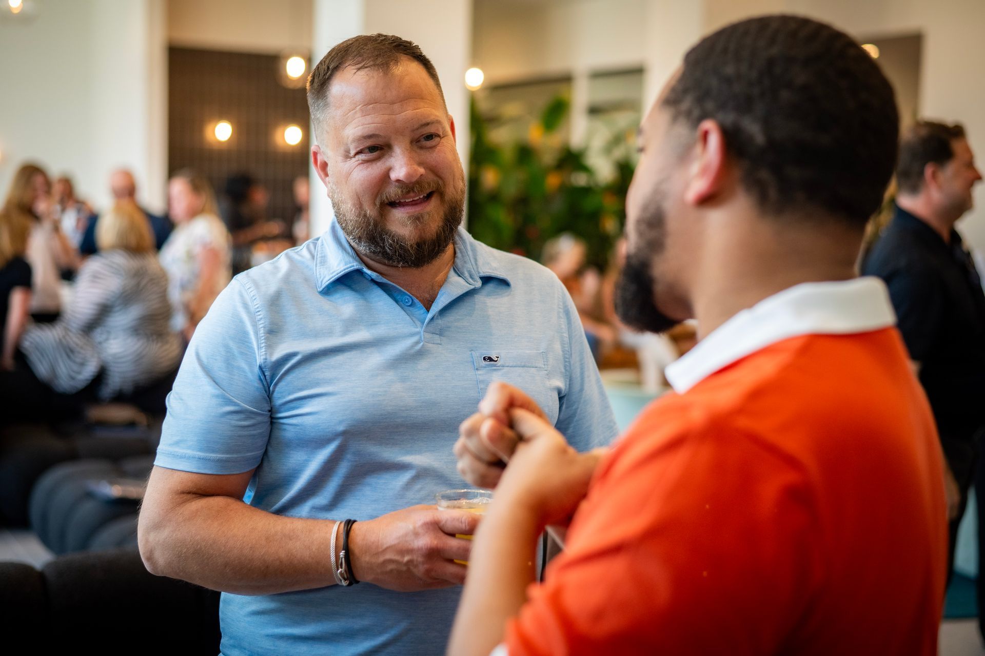 Two men in conversation at a social gathering. One in a blue polo, the other in an orange shirt.