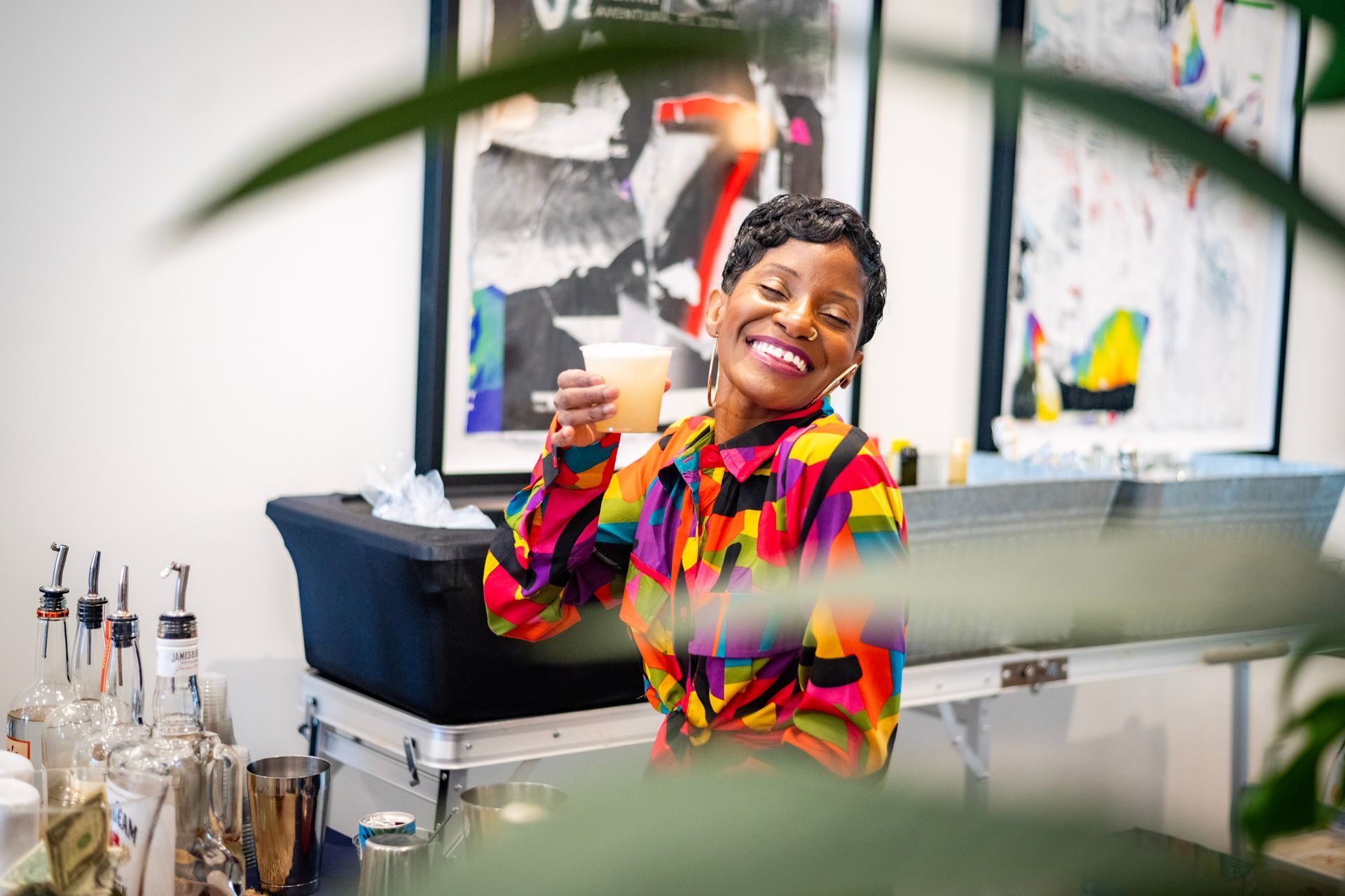 Woman smiling, holding a drink, behind a bar with art in the background. She wears a colorful shirt.