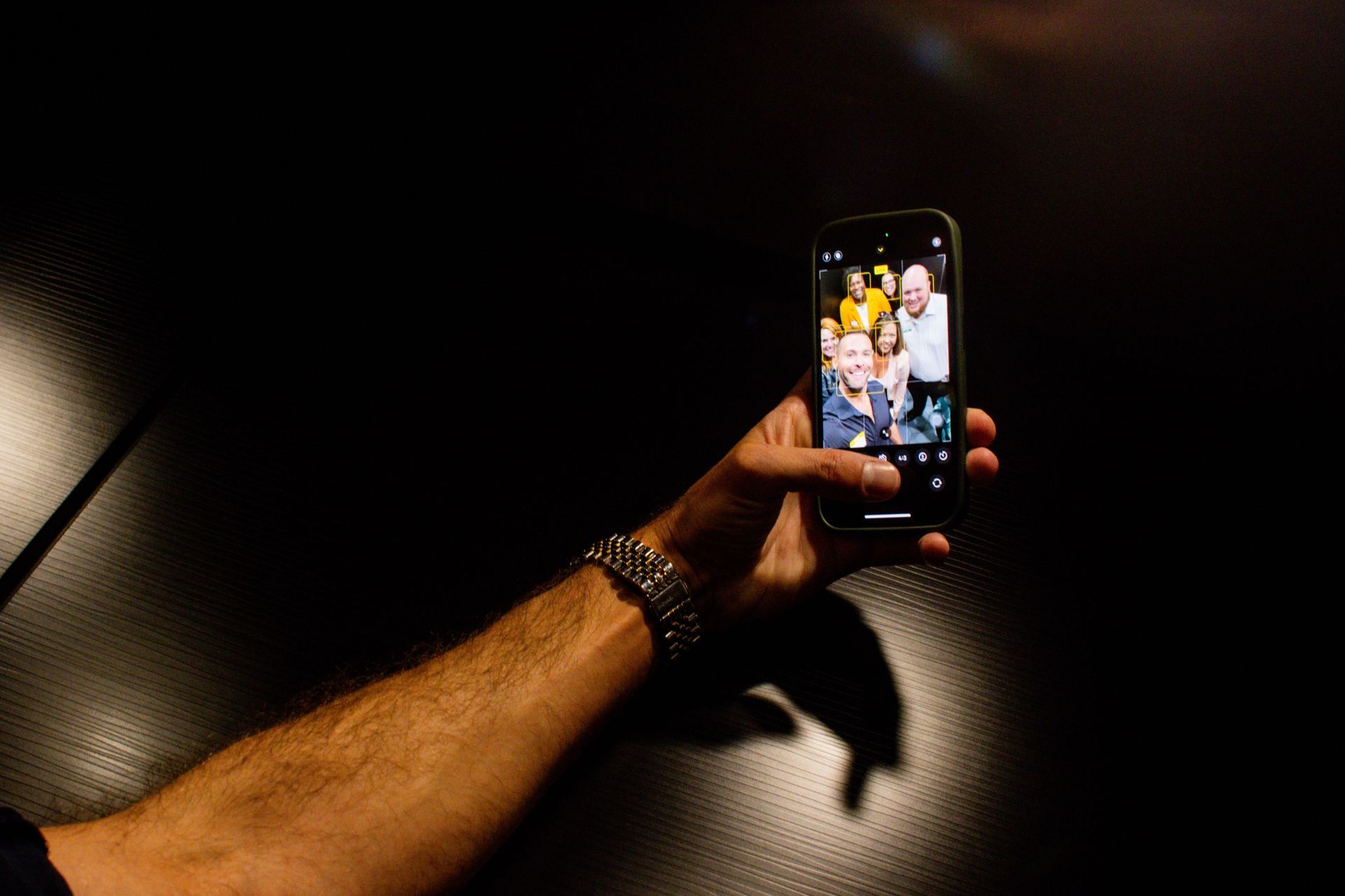 Person's arm holding smartphone, taking selfie with smiling people. Dark background, watch on wrist.