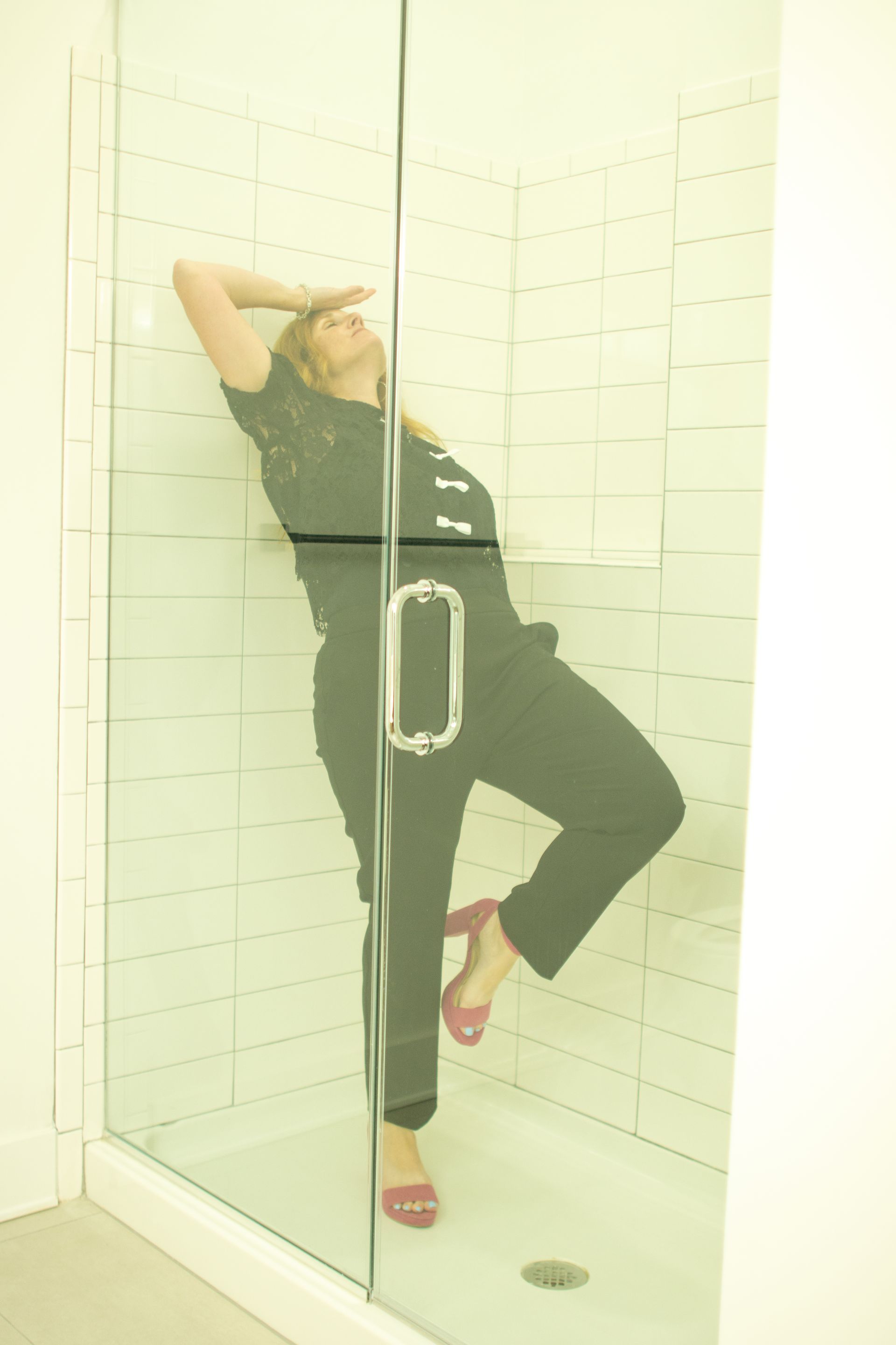 Woman in black jumpsuit and pink heels in shower, leaning against glass, shielding eyes.