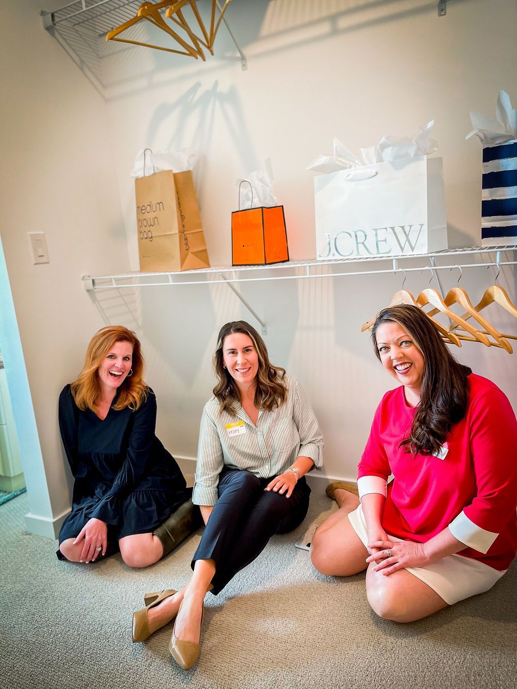 Three women sit in a closet, smiling. Shopping bags on shelf above.