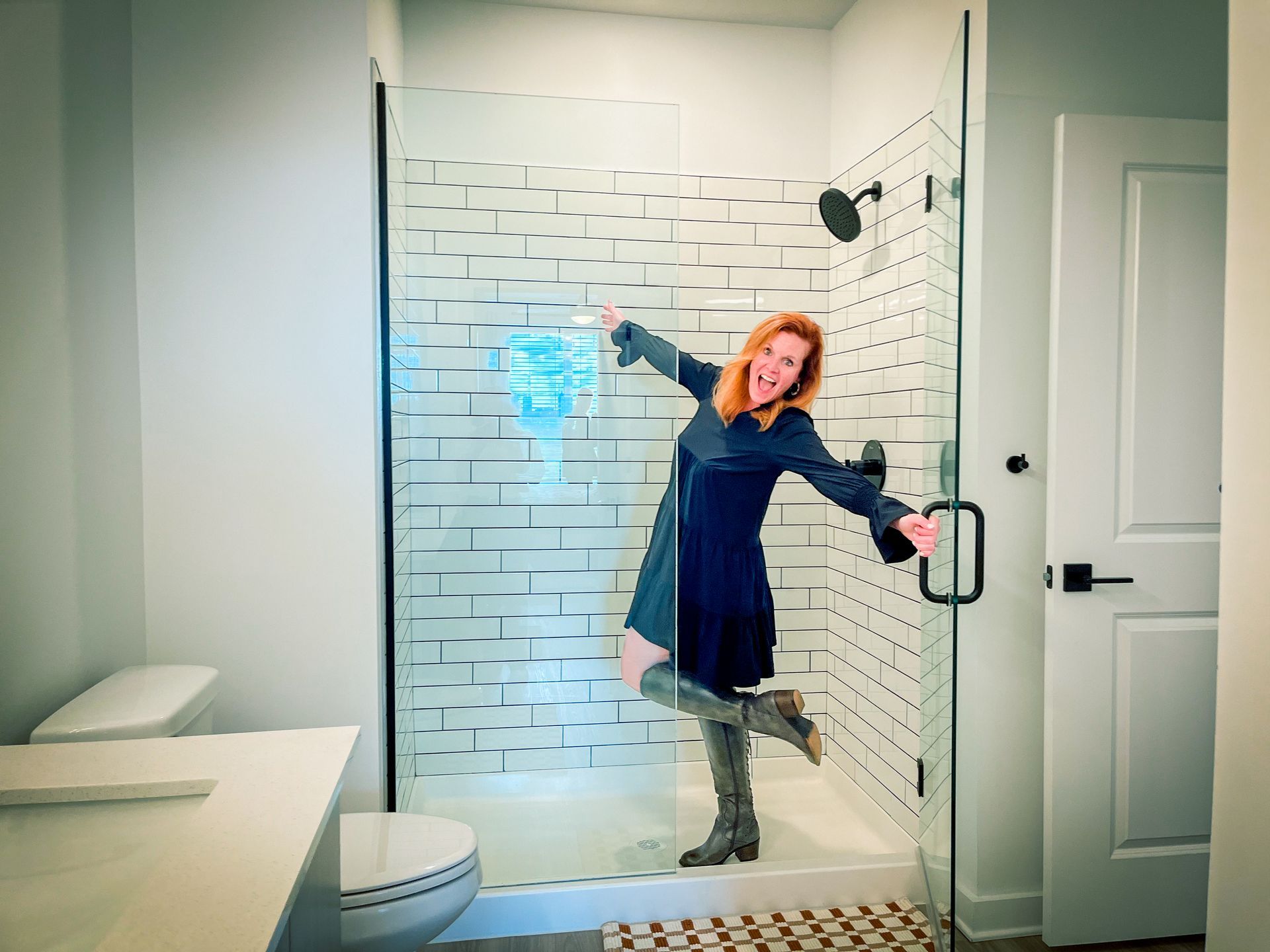 Woman in a dark dress and boots dances in a shower with white brick-like tiles, a white toilet, and a black showerhead.