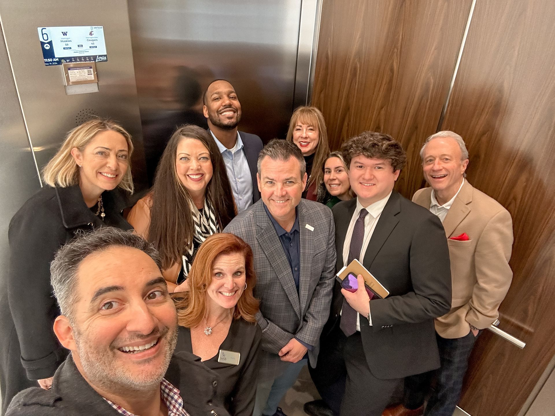 Group of people in elevator, taking a selfie. Smiling faces, various attire. Wooden paneling.